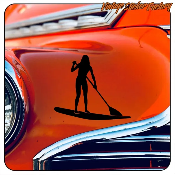 Silhouette noire d’une femme avec planche et pagaie posée sur la carrosserie orange d’une voiture classique