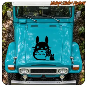 Adhesivo de Totoro y dos figuras pequeñas aplicado sobre el capó de un vehículo turquesa